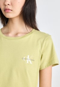 Šviesiai žalia medvilnės marškinėliai su trumpomis rankovėmis, turintys baltą "CK" logotipą ir tekstą "Calvin Klein Jeans". Lygi tekstūra, apvalus kaklas.