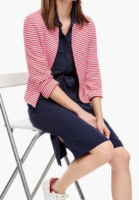 Gestreepte rood-witte cardigan over een marineblauwe jurk, met opgerolde mouwen en zijsplitten, gecombineerd met witte sneakers met rode accenten.