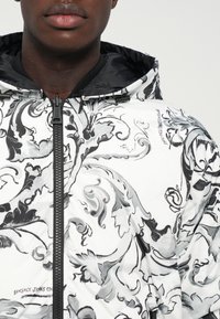 Tuvumā redzams cilvēks, kas valkā baltu jaku ar melniem un pelēkiem ornate ziedu rakstiem un melnu rāvējslēdzēju, redzama arī kapuces oderes daļa.