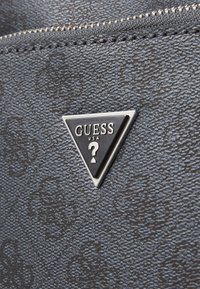 Juoda sintetinio pluošto medžiaga su tekstūru, su trikampiu sidabriniu emblemu, ant kurios yra "GUESS USA" ir klausimo ženklas.
