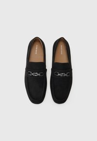 Mocasines de ante negro con una cadena metálica decorativa y un detalle trenzado a lo largo del empeine. Textura suave con suelas planas y diseño minimalista.