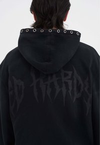 Sudadera negra con un gran gráfico en la parte posterior que presenta letras estilizadas. Ojales de metal bordean el borde de la capucha. Textura suave.