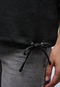 Maglione nero a coste con cintura annodata e occhielli argentati, abbinato a jeans grigi. La texture è evidente, mostrando una finitura in tessuto morbido.