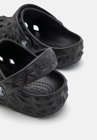 Sabots Crocs noirs avec un motif géométrique texturé et des sangles arrière réglables, présentés en gros plan sur un fond blanc.