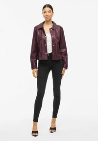 Veste en simili cuir bourgogne avec un col, boutons-pression à l'avant et deux poches, portée sur un haut blanc et un jean skinny noir.