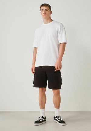 Junger Mann steht, trägt ein schlichtes weißes übergroßes T-Shirt, schwarze Cargo-Shorts, weiße Sportsocken und schwarze Turnschuhe mit weißen Schnürsenkeln.