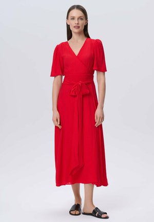 Abito rosso avvolgente con maniche a sbuffo corte, una cintura in vita e tessuto fluido. Indossato con sandali neri flat. Texture liscia, design a tutta lunghezza.