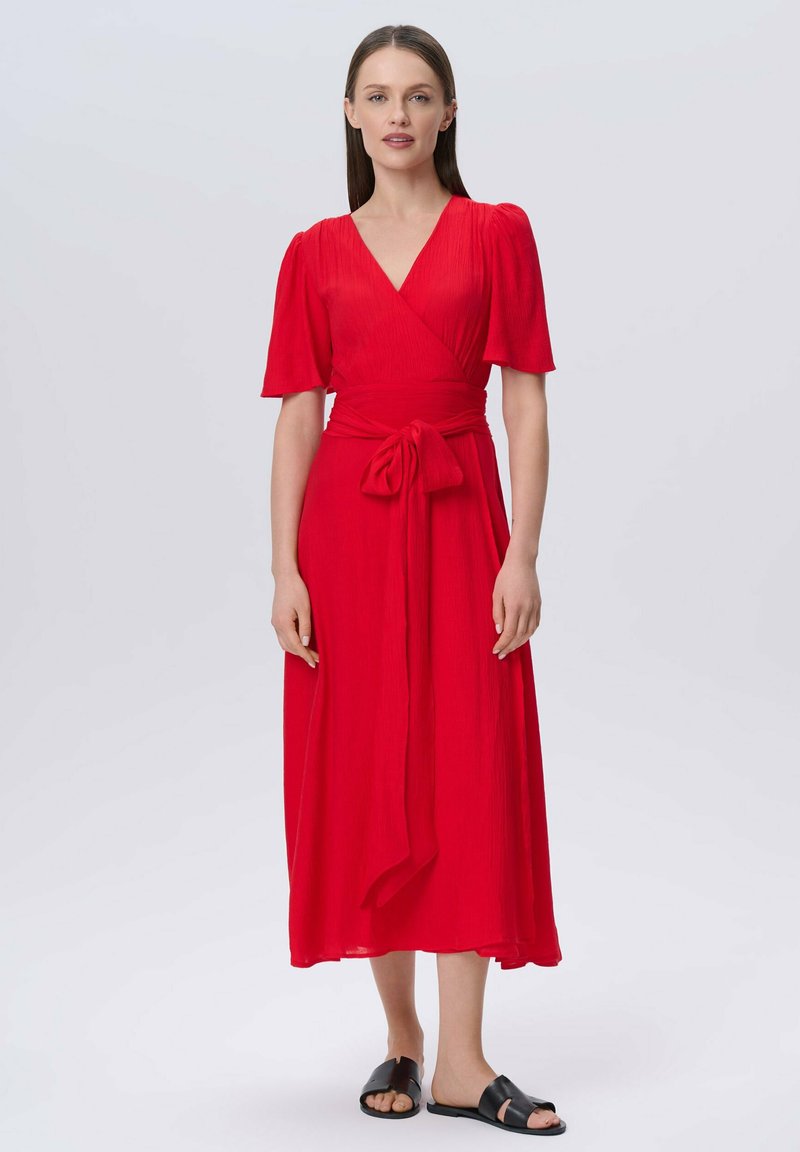Rotes Wickelkleid mit kurzen Puffärmeln, einem Taillengürtel und fließendem Stoff. Getragen mit schwarzen flachen Sandalen. Glatte Textur, bodenlanges Design.