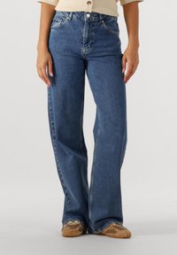 Nő kék magas derekú, bőszárú farmernadrágot és bézs sportcipőt visel, és egy egyszerű világos háttér előtt áll.