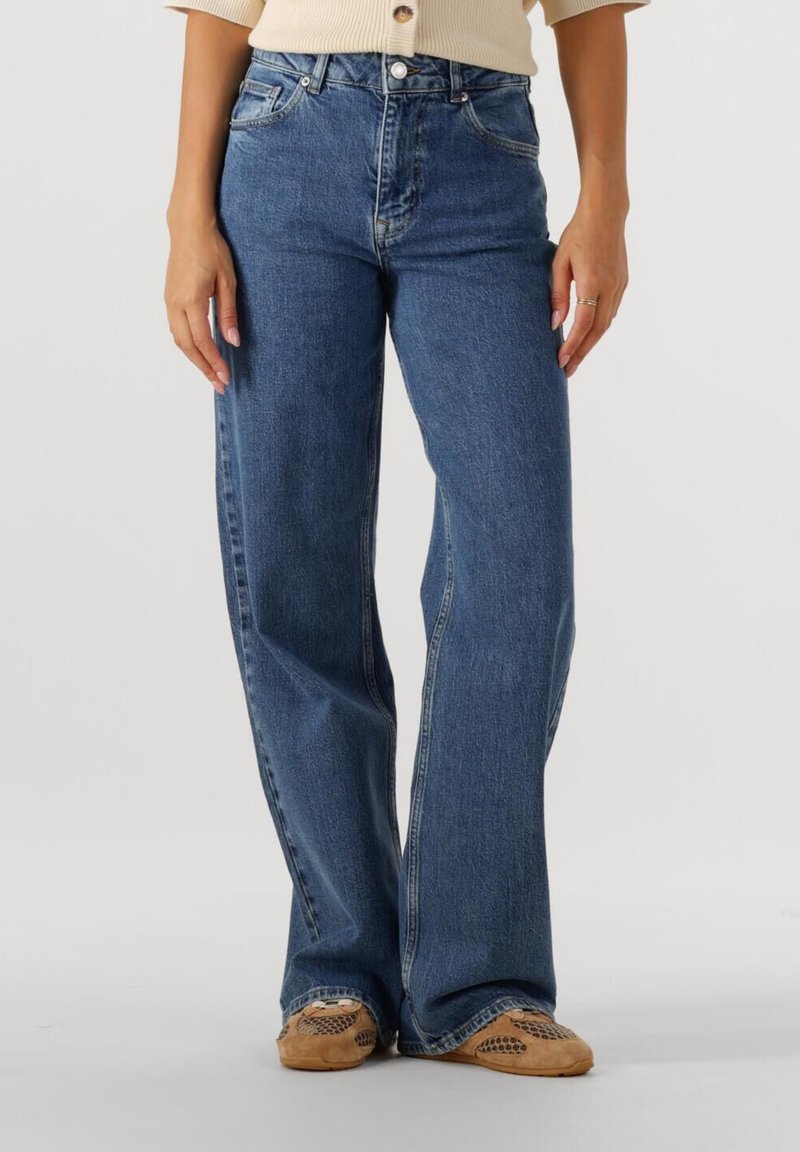 Nő kék magas derekú, bőszárú farmernadrágot és bézs sportcipőt visel, és egy egyszerű világos háttér előtt áll.