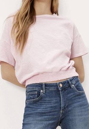 Kvinde iført en lyserød T-shirt med korte ærmer og elastisk kant, sat sammen med blå denimjeans, stående foran en ensfarvet baggrund.