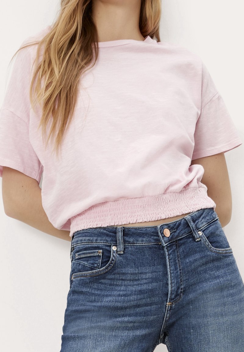 Femme portant un haut rose clair à manches courtes avec ourlet élastique, associé à un jean en denim bleu, debout devant un fond uni.