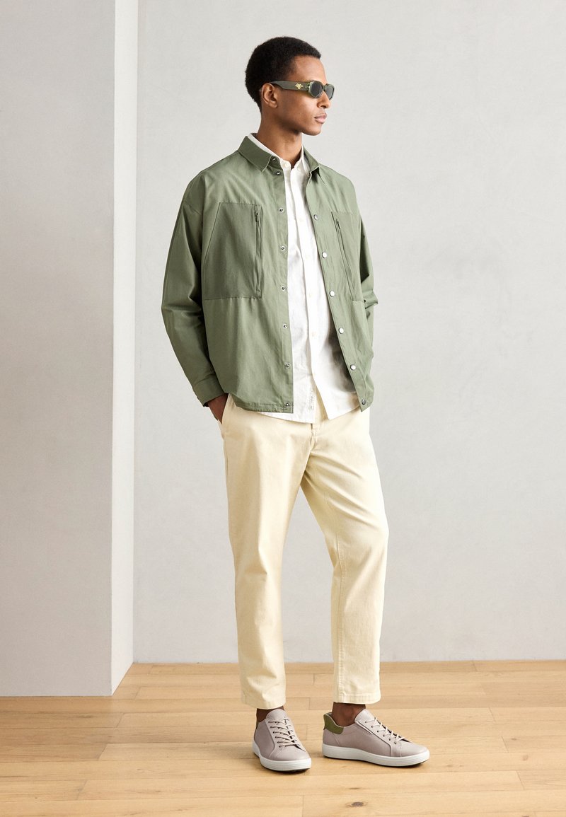 Hellgrüne leichte Jacke, weißes Hemd mit Knöpfen, beigefarbene Chinos und graue Sneaker mit grünen Akzenten. Modell steht in einem minimalistischen Raum.