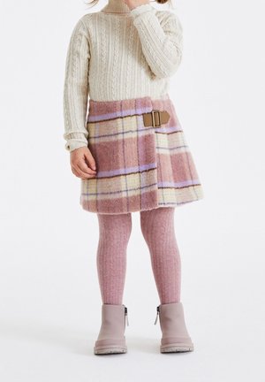 Barn iført cremefarvet kabelstrikket sweater, lyserødt og cremefarvet ternet nederdel med spænde, lyserøde strukturerede strømpebukser og lyserøde støvler, stående mod en hvid baggrund.