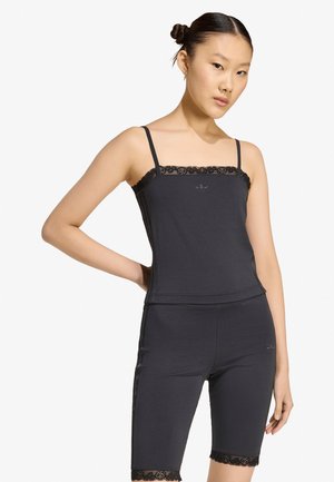 Femme aux cheveux foncés en chignons, portant un top à bretelles fines orné de dentelle noire et un short assorti à la longueur du genou, debout, une main sur la hanche.