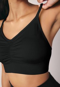 Zwart mouwloos bralette met een geplooide voorkant, gladde stof en geribbelde onderband. Verstelbare banden zijn zichtbaar op één schouder.