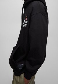 Osoba ubrana w czarną bluzę z kapturem z przednią kieszenią i napisem z różową grafiką dłoni na piersi, w połączeniu z beżowymi spodniami.