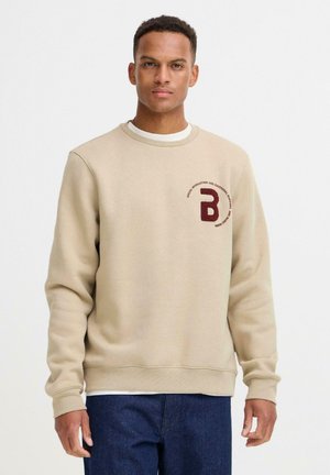 Sweatshirt beige en tissu doux, avec un col rond et des poignets côtelés. Un logo bordeaux foncé est brodé sur la poitrine.