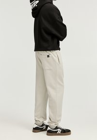 Schwarzer cropped Hoodie mit weißem Logo, kombiniert mit hellgrauen Sweatpants mit seitlichen Taschen und schwarzen Sneakers mit weißen und braunen Akzenten.