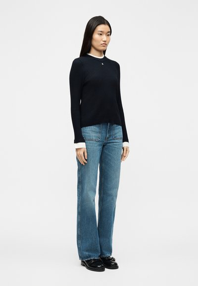 Γυναίκα με μακριά σκούρα μαλλιά που φοράει μαύρο ριμπ πουλόβερ πάνω από λευκές μαν σέτες, μπλε τζιν ευθείας γραμμής και μαύρα μοκασίνια, στέκεται μπροστά από απλό φόντο.