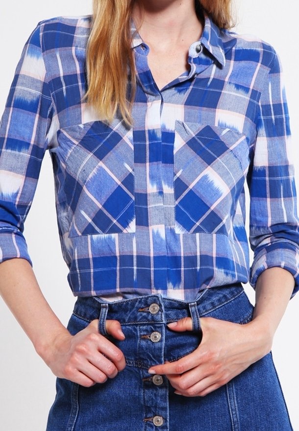 Chemise à carreaux bleue à boutons avec deux poches poitrine, présentant un mélange de tons bleu clair et foncé, des accents rose pâle, et des manches retroussées.