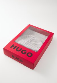 White knitted beanie displayed in a transparent red box. The box features bold black "HUGO" branding on the front.