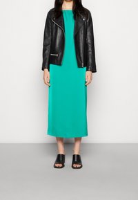 Schwarze Lederbikerjacke über einem einfarbigen türkisfarbenen Maxikleid. Das Modell trägt schwarze offene Sandalen. Glatte Textur, knielanges Kleid, lässiger Stil.