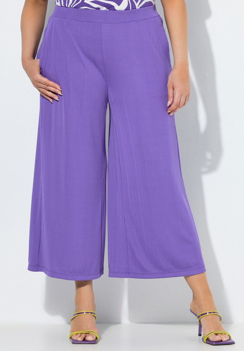 Femme portant un pantalon violet taille haute à jambes larges, assorti à des sandales à talons jaunes à brides, debout sur un fond blanc.