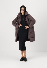 Bruine pufferjack met capuchon, met een geborduurd ontwerp en zijzakken, gedragen over een zwarte geribbelde jurk en zwarte schoenen.