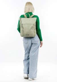 Zaino in pelle verde di forma quadrata, con manici superiori e tasca con zip frontale, abbinato a jeans a gamba larga di colore blu chiaro e scarpe bianche.