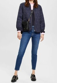 Chaqueta bomber acolchada azul marino, camisa rosa claro, suéter azul marino, vaqueros azules y mocasines negros. Pequeño bolso cruzado negro con detalles dorados.