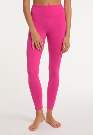 Vrouw die felroze strakke leggings draagt, blootsvoets staand tegen een effen lichte achtergrond.