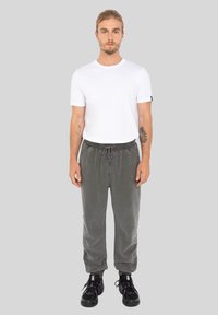 Mężczyzna z tatuażami ubrany w biały t-shirt, szare dresy i czarne sneakersy stoi na jednolitym szarym tle.