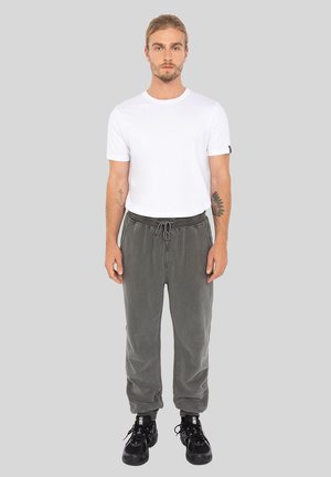 Uomo con tatuaggi che indossa una maglietta bianca, pantaloni della tuta grigi e sneakers nere, in piedi davanti a uno sfondo grigio uniforme.