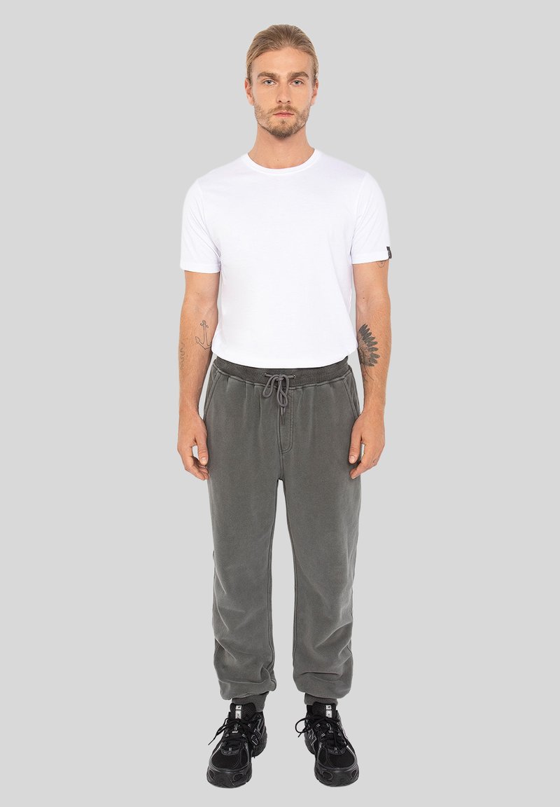 Mężczyzna z tatuażami ubrany w biały t-shirt, szare dresy i czarne sneakersy stoi na jednolitym szarym tle.