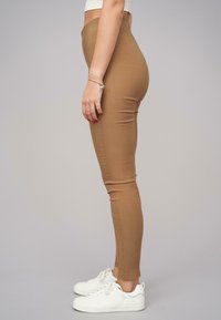Barna, testhezálló leggings, rugalmas anyagból készült, magas derékkal és sima textúrával. Fehér sportcipővel párosítva.