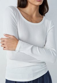 Maglia bianca a maniche lunghe a coste con scollatura rotonda, caratterizzata da una silhouette aderente e da una texture morbida, abbinata a pantaloni scuri a righe verticali.