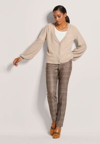 Beige Strickjacke mit tiefem V-Ausschnitt und Knopfleiste, kombiniert mit braunen Karohosen und braunen Absatzschuhen mit Kettendetail.