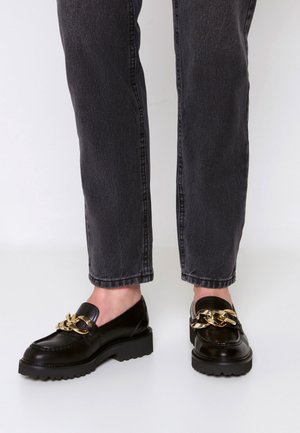 Sorte læderloafers med kraftige såler og stor guldkæde detalje, båret med mørkegrå jeans med lige ben.