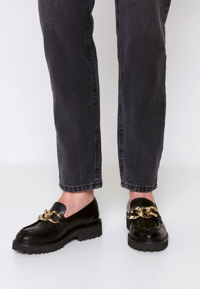 Mocassins en cuir noir avec semelles épaisses et grand détail chaîne en or, portés avec un jean droit gris foncé.