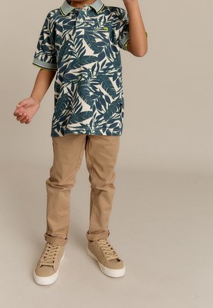 Barn iført beige bukser, brune sneakers og en grøn polo med bladmønster og korte ærmer, stående på en ensfarvet baggrund.