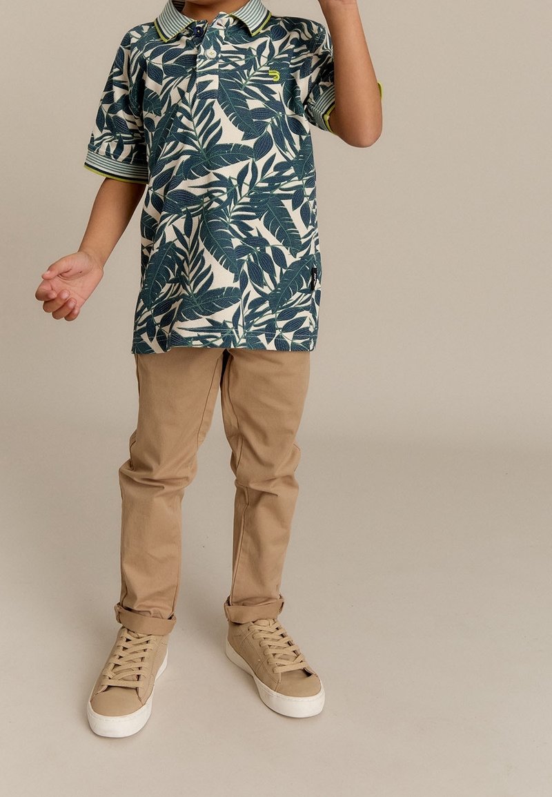 Enfant portant un pantalon beige, des baskets marron clair et un polo vert à motif de feuilles à manches courtes, debout sur un fond uni.