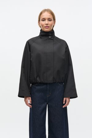 Veste noire courte avec col montant, fermeture à boutons et manches larges. Le tissu semble lisse et résistant aux intempéries. Portée avec un jean foncé.