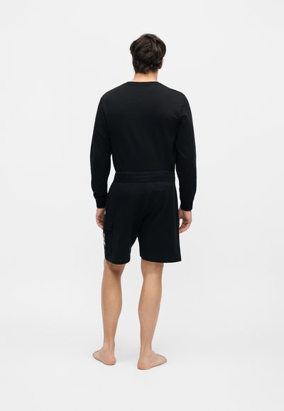 Άνδρας στέκεται ξυπόλητος με την πλάτη γυρισμένη, φοράει μαύρο μακρυμάνικο πουκάμισο και μαύρες βερμούδες μέχρι τα γόνατα με πλαϊνή τσέπη.