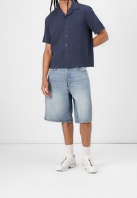 Camisa de manga corta de botones color navy, pantalones cortos de mezclilla azul claro hasta la rodilla, zapatillas blancas con acentos multicolores, sobre un fondo liso.