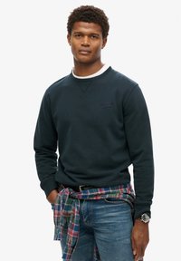 Sudadera azul marino con cuello redondo y puños acanalados, que presenta un pequeño logo. Llevada con una camisa de cuadros atada alrededor de la cintura y jeans.
