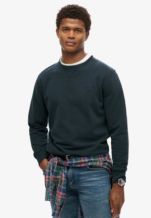 Sudadera azul marino con cuello redondo y puños acanalados, que presenta un pequeño logo. Llevada con una camisa de cuadros atada alrededor de la cintura y jeans.