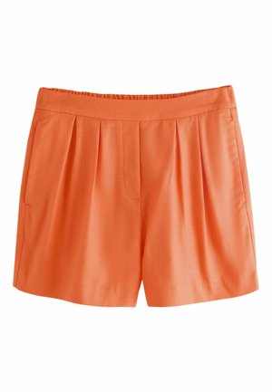 Kvinde iført lyse orange skræddersyede shorts med baglommer, står indendørs med hænderne delvist i sidelommerne.