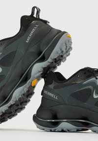 Schwarze Sportschuhe mit einem Obermaterial aus Mesh, robusten Gummisohlen und einer grauen Zwischensohle. Mit "Merrell"-Markenlogo und einer Schlaufe an der Ferse.