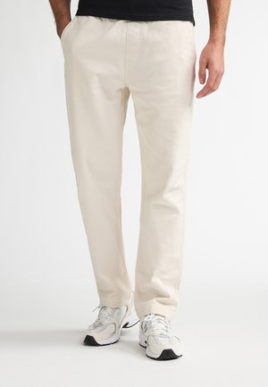 Pantalones de algodón beige con un corte holgado, con bolsillos laterales y una textura suave, combinados con zapatillas deportivas de color gris claro.